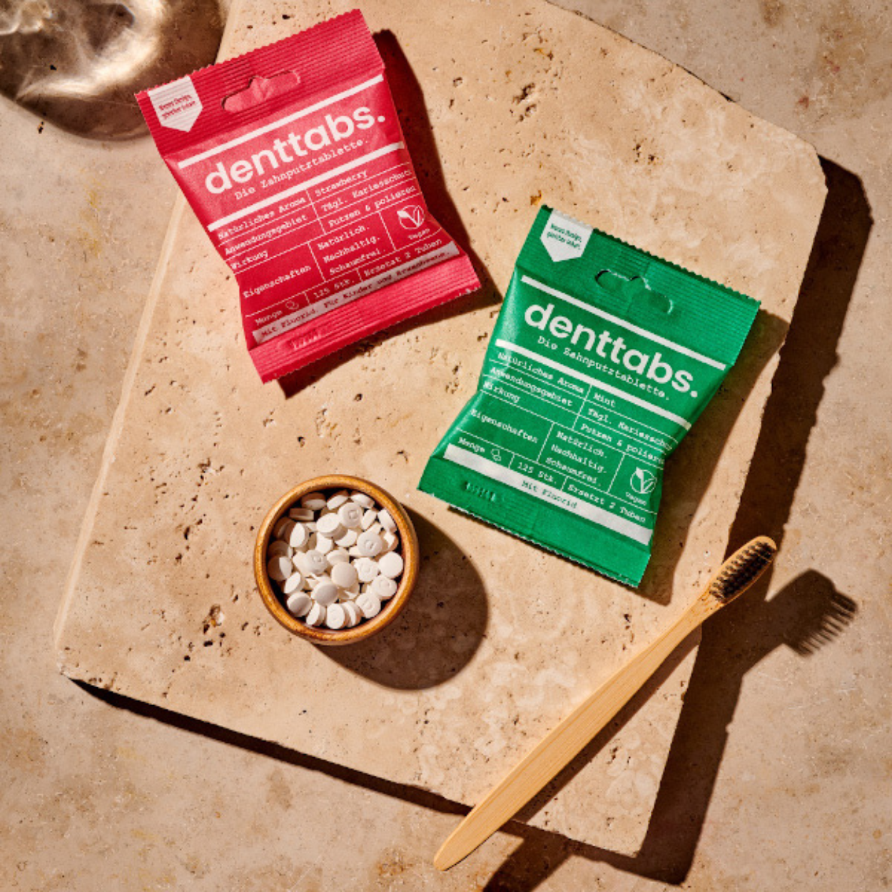Denttabs toothpaste tablets in red and green sachets on a beige stone surface, with a small bowl of white tablets and a bamboo toothbrush.